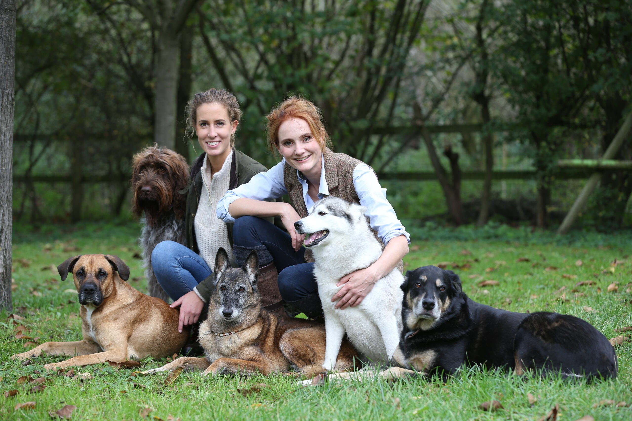 Ramona Teschner und Julia Appelt sitzen mit ihren Hunden auf einer Wiese und lächeln freundlich in die Kamera.